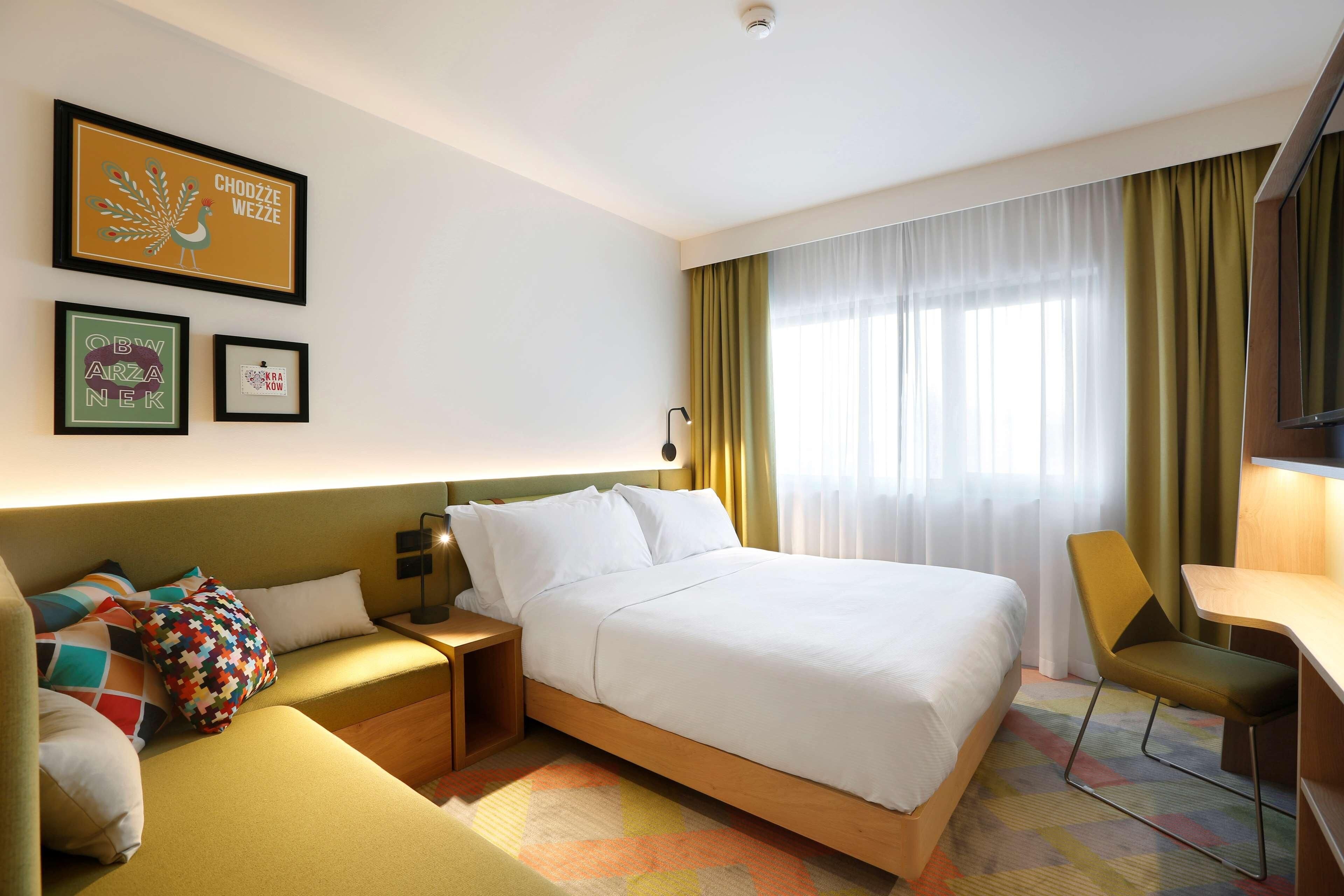 A hotel room with a bed, a desk, a chair, a nightstand, and a lamp. There are curtains on the window and a wall-mounted light fixture.