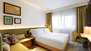 A hotel room with a bed, a desk, a chair, a nightstand, and a lamp. There are curtains on the window and a wall-mounted light fixture.