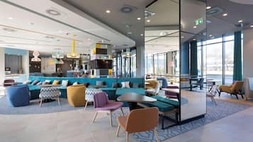 A modern lounge area with seating arrangements, a kitchen in the background, and large windows providing natural light.