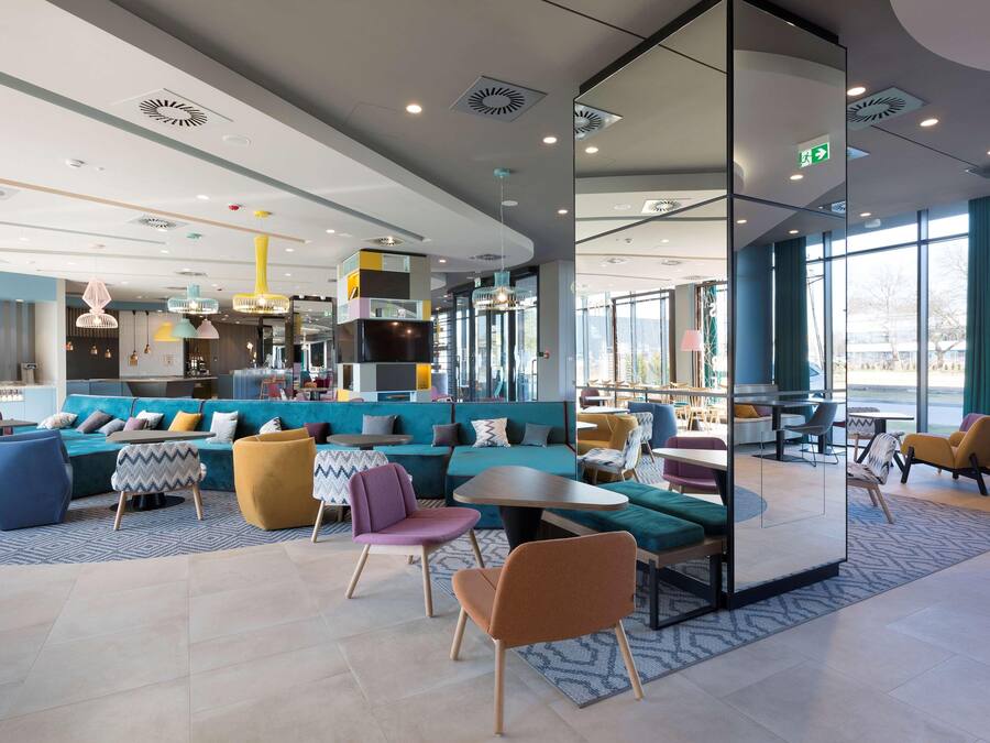 Un moderno espacio de salón con arreglos de asientos, una cocina al fondo y grandes ventanales que ofrecen luz natural.
