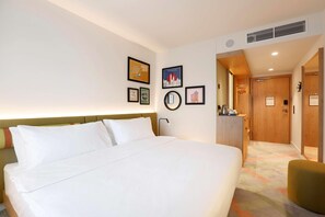A hotel room with a large bed, a desk, and a chair. There are framed pictures on the wall and a door leading to another room. - Hampton By Hilton Krakow Airport (Zabierzow)