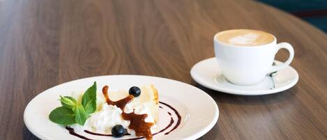 Un plato de postre con crema batida, arándanos y salsa de chocolate sobre un plato blanco, acompañado por una taza de café sobre su platillo.