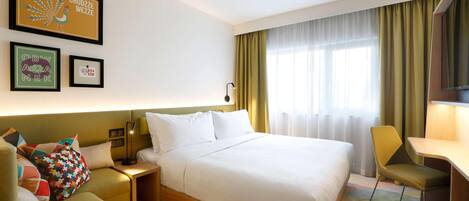 A hotel room with a bed, a desk, a chair, a nightstand, and a wall with framed artwork.