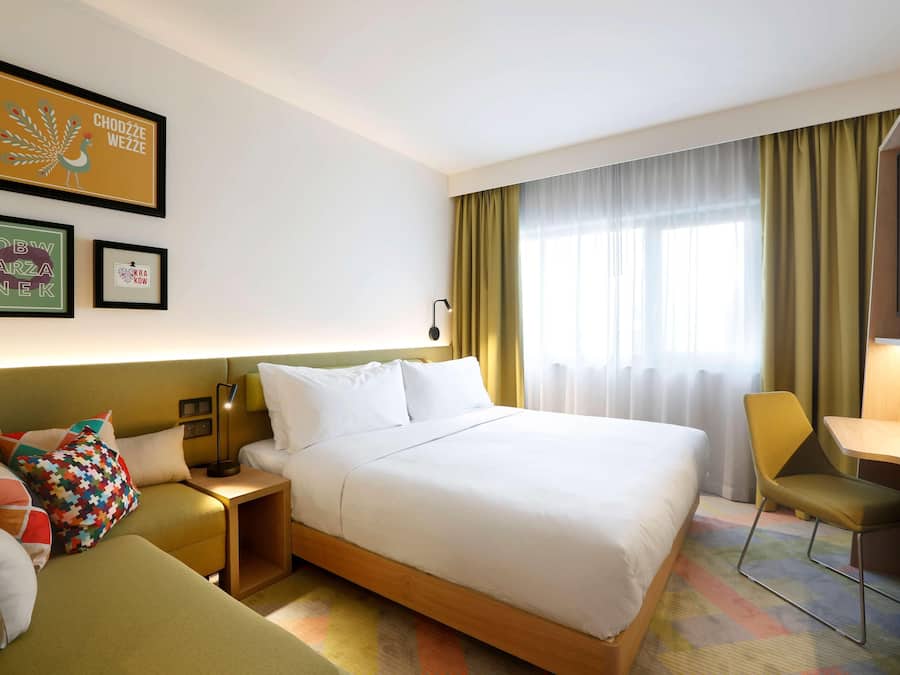 A hotel room with a bed, a desk, a chair, a nightstand, and a wall with framed artwork.
