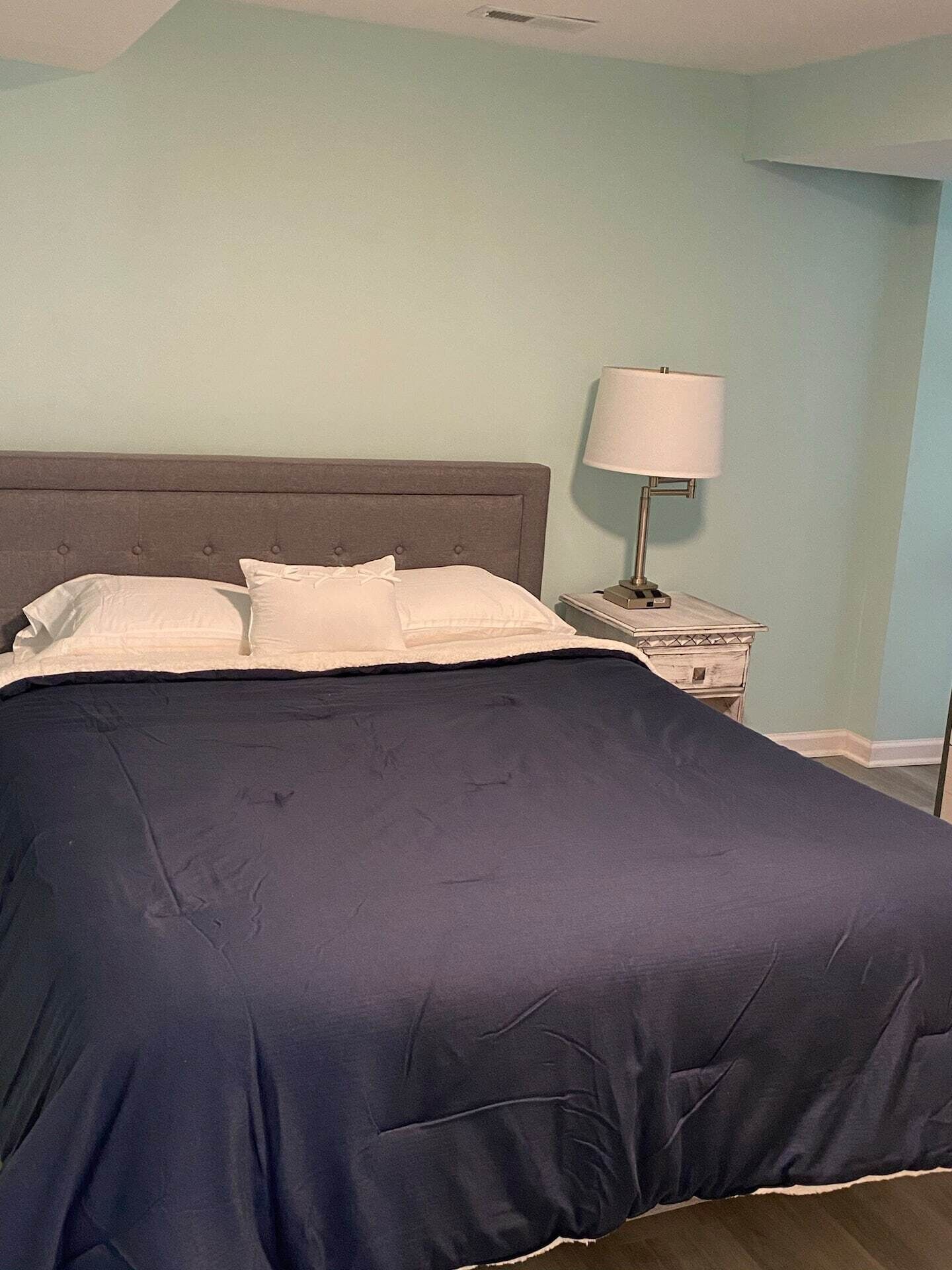 Ein ordentlich bezogenes Bett mit dunkelblauer Bettdecke und weißen Kissen, ein Nachttisch mit Lampe sowie eine hellblaue Wand.