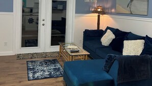 Una sala de estar con ventilador de techo, un sofá azul, una mesa de centro de vidrio y un cuadro de un pájaro en la pared.
