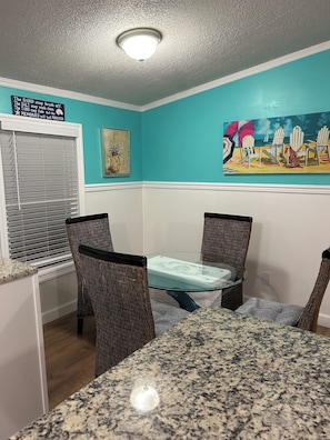 Relax in this inviting dining area with a round table and wicker chairs, surrounded by a beach-themed painting.