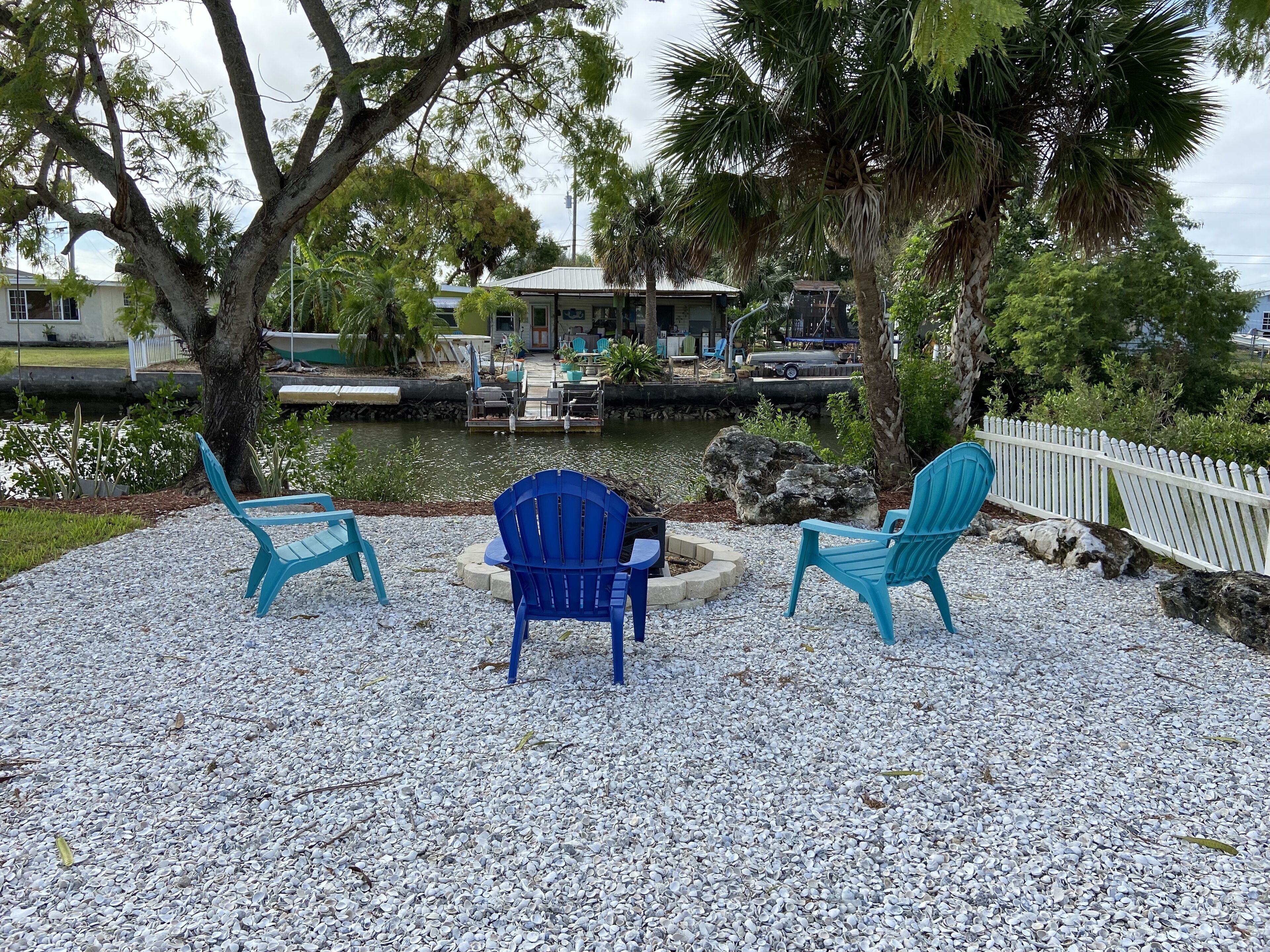A patio area with three blue Adirondack chairs, a small table, and a pathway leading to a dock over water.