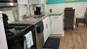 Una cocina con gabinetes blancos, una estufa negra y una cafetera. Hay un cuadro en la pared y una silla en el comedor.