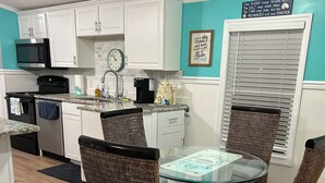 Una cocina compacta con armarios blancos, microondas y cafetera. Hay un comedor con sillas de mimbre y una mesa de vidrio.