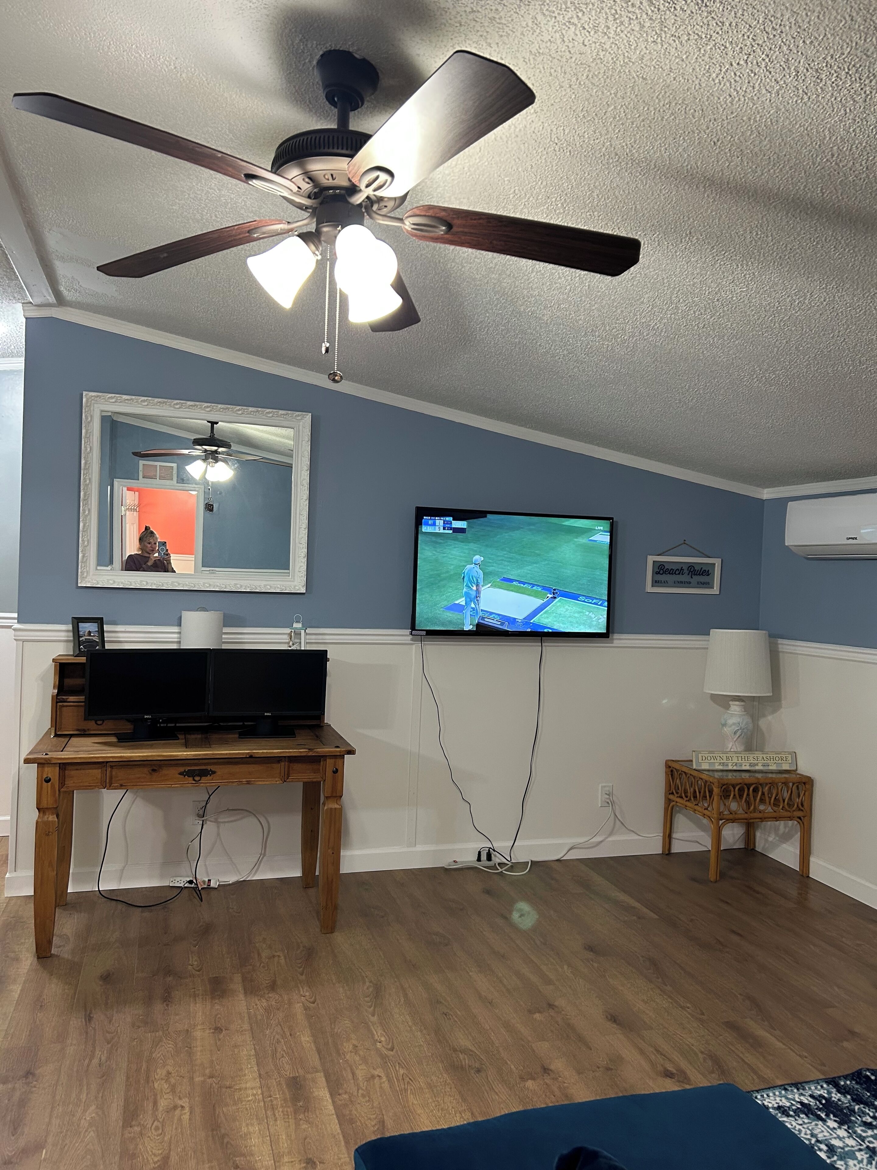 A room with a ceiling fan, a TV mounted on the wall, a wooden desk, and a small table with a lamp.