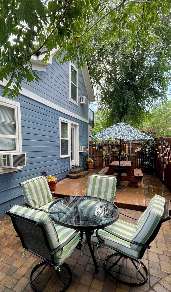 Un patio con una mesa de vidrio rodeada de sillas a rayas, una terraza de madera y una casa azul con contraventanas blancas.