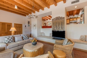 A spacious living room with a large sofa, a coffee table, and a television. There is a kitchen area with wooden cabinets and a refrigerator. - Acoma - Majestic Views, Near the Santa Fe Opera and Four Seasons in Tesuque (Santa Fe)