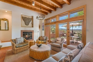 A spacious living room with a fireplace, wicker furniture, and large windows offering a view of the outdoors. - Acoma - Majestic Views, Near the Santa Fe Opera and Four Seasons in Tesuque (Santa Fe)