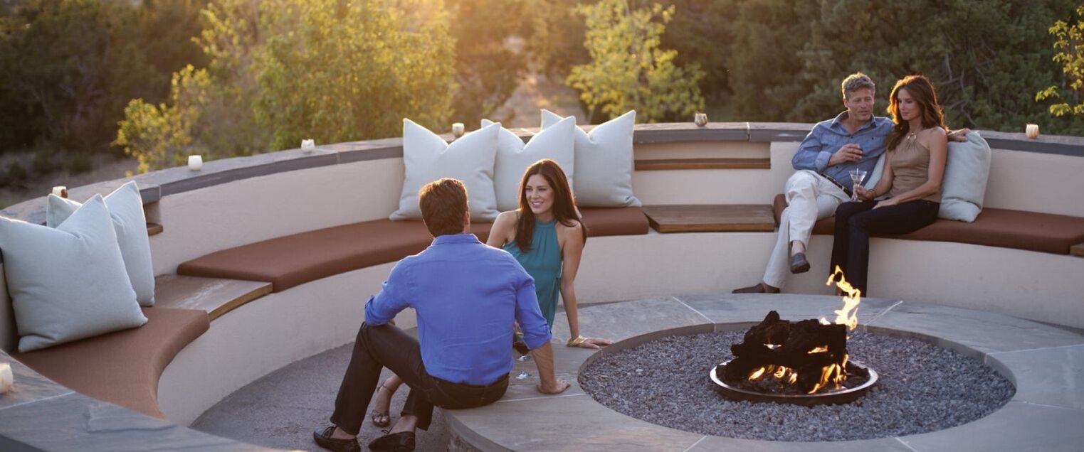 A circular outdoor seating area with a fire pit, cushioned benches, and a stone floor.