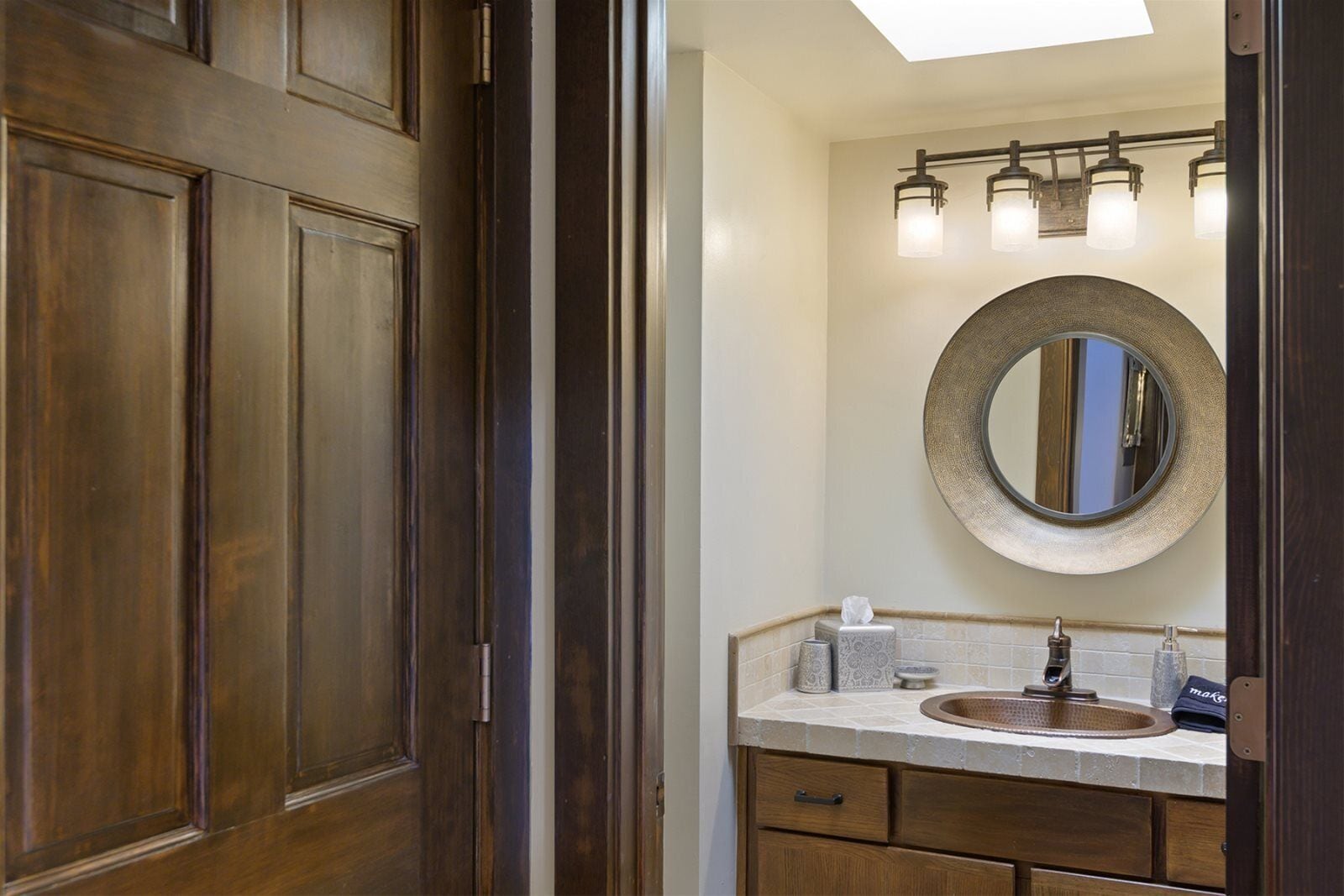 A bathroom with a wooden door, a round mirror, a sink with a towel, and a light fixture above the mirror.