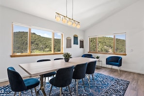 A modern dining room with a large table, blue chairs, and a view of a forested hill.