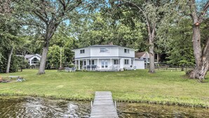 A two-story house with a covered porch, a wooden dock extending into a pond, and a grassy yard with trees. - Love The 8 Lakes Life! (Pinckney)