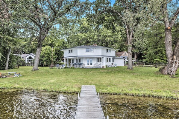 A two-story house with a covered porch, a wooden dock extending into a pond, and a grassy yard with trees. - Love The 8 Lakes Life! (Pinckney)