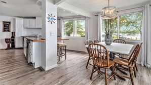 A modern kitchen with white cabinetry, a wooden dining table with chairs, and a large window with a view of trees. - Love The 8 Lakes Life! (Pinckney)