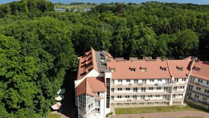 A multi-story hotel with a red-tiled roof, surrounded by dense green forest, and a view of the sea in the background.