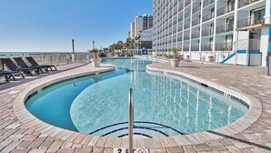 Piscina com design curvo, rodeada por espreguiçadeiras e um edifício com várias varandas.