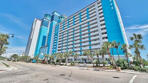 Um hotel alto com fachada azul e branca, vários andares e um pátio central com palmeiras.