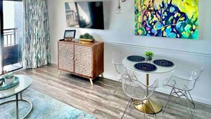 Uma sala de estar moderna com uma mesa de jantar redonda, cadeiras transparentes, um armário de madeira e uma pintura abstrata colorida na parede.
