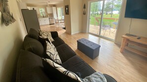 A spacious living room with a dark-colored sofa, a small ottoman, and a television on a wooden stand. A sliding glass door leads to a patio with outdoor furniture. - Lakeside Cottage Just Off Snowmobile/ATV Trail! (Lake Linden)