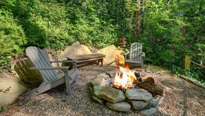 A forested outdoor seating area with Adirondack chairs, a wooden table, and a stone fire pit.