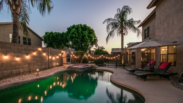 A swimming pool with a hot tub, surrounded by a brick wall and palm trees, with lounge chairs and string lights. - Sunshine and Good Times! Massive 5/bdrm with Pool, Hot Tub, and Game Room!  (Gilbert)
