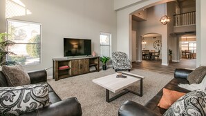 A spacious living room with a flat-screen TV, leather sofas, a coffee table, and a dining area in the background. - Sunshine and Good Times! Massive 5/bdrm with Pool, Hot Tub, and Game Room!  (Gilbert)