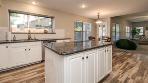 A spacious kitchen with white cabinetry, granite countertops, and a large island. There is a dining area with a chair and a table, and a green bean bag chair in the living room. - Sunshine and Good Times! Massive 5/bdrm with Pool, Hot Tub, and Game Room!  (Gilbert)