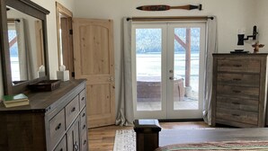 Un dormitorio con cómoda de madera, cama con manta estampada, un espejo grande y puerta corrediza de vidrio que da a un balcón con vista al lago.