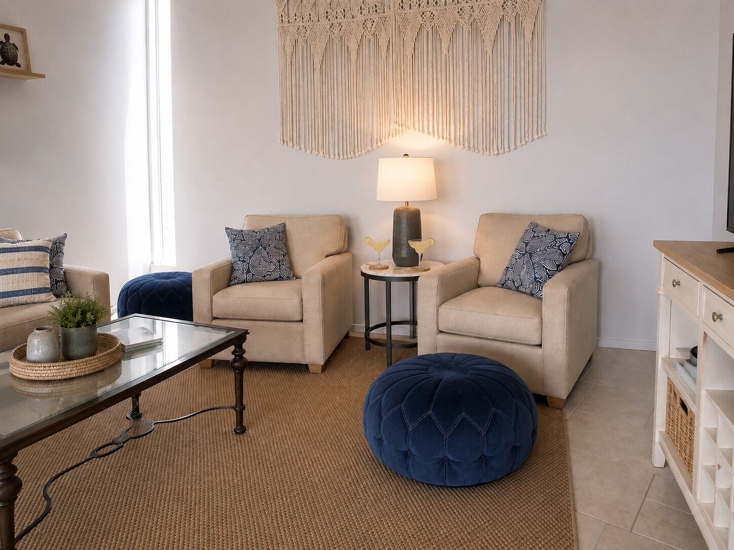 Un salon comprenant deux fauteuils, une table basse, une table d’appoint avec une lampe, un pouf bleu et une décoration murale en macramé.