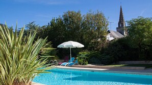 En swimmingpool med solstolar och ett parasoll, omgiven av grönska och en kyrka i bakgrunden.