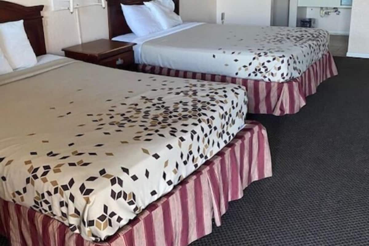 Ein Hotelzimmer mit zwei Betten, jeweils mit einem gemusterten Überwurf und einem gestreiften Bettrock. Es gibt einen Nachttisch mit einer Lampe, ein Bild an der Wand und ein durchgegangenes Badezimmer ist sichtbar.