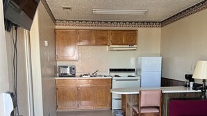 A compact kitchen with wooden cabinets, a white stove, a sink, and a small dining area with a chair and table. - Hallmark Inn and Suites (Killeen)