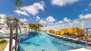 A swimming pool with a curved edge, surrounded by palm trees and plants, with a clear blue sky in the background. - Apart térreo no In Mare Bali Cotovelo (Parnamirim)
