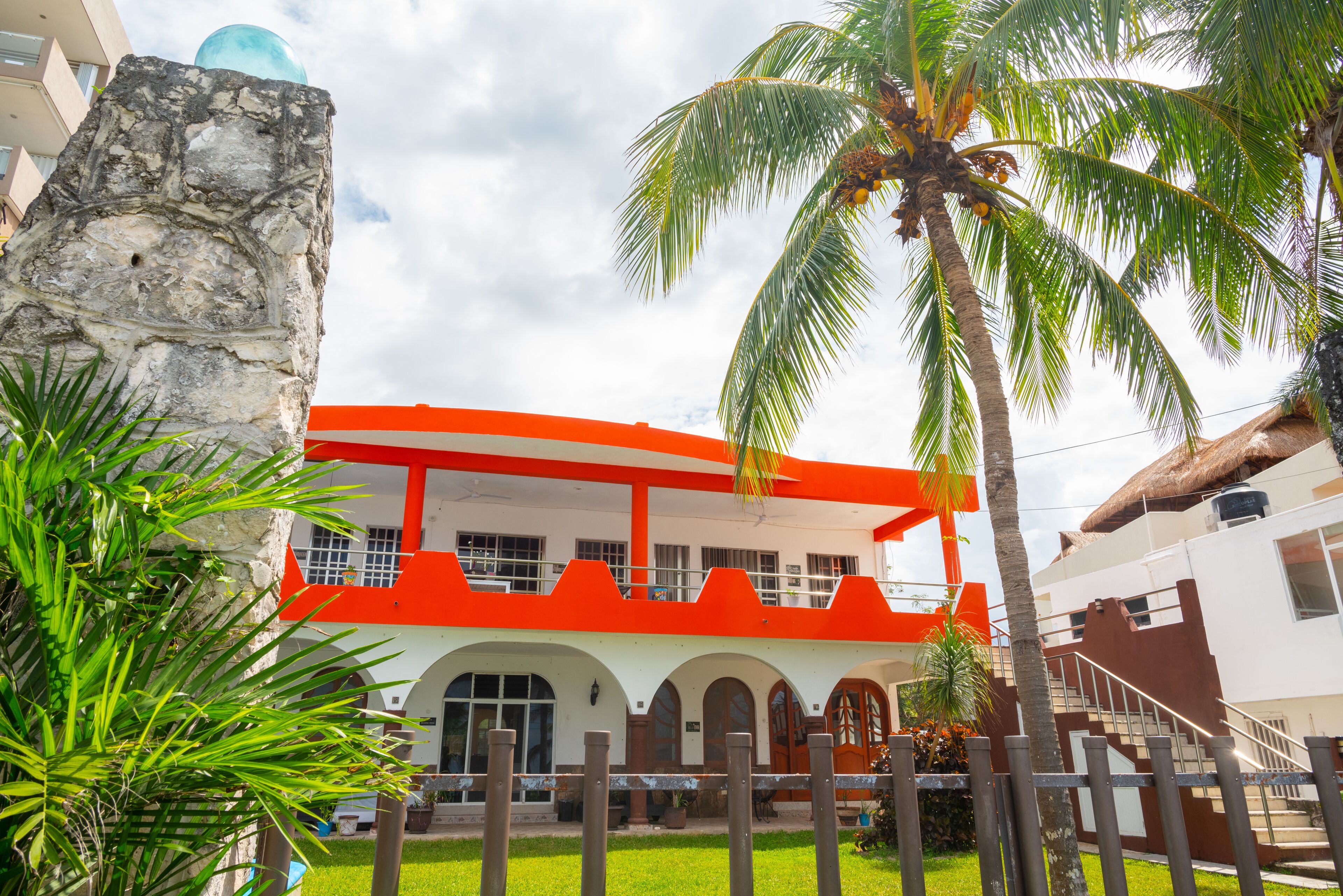 A building with a white facade and orange accents, featuring a stone structure to the left and a palm tree in the foreground.
