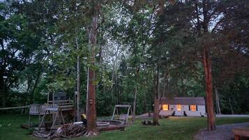 A rustic cabin surrounded by dense trees, with a swing set and a wooden deck.