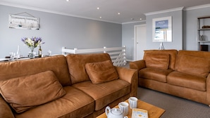 A modern living room with two brown leather sofas, a coffee table with a teapot and mugs, a dining table set with glasses and a vase of flowers, and a white railing in the background.