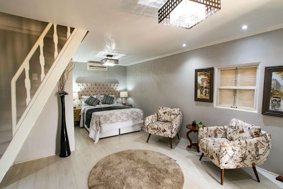 Une chambre à coucher comprenant un lit, des tables de chevet, une chaise, un tapis rond, un escalier et une fenêtre décorée d’œuvres d’art.