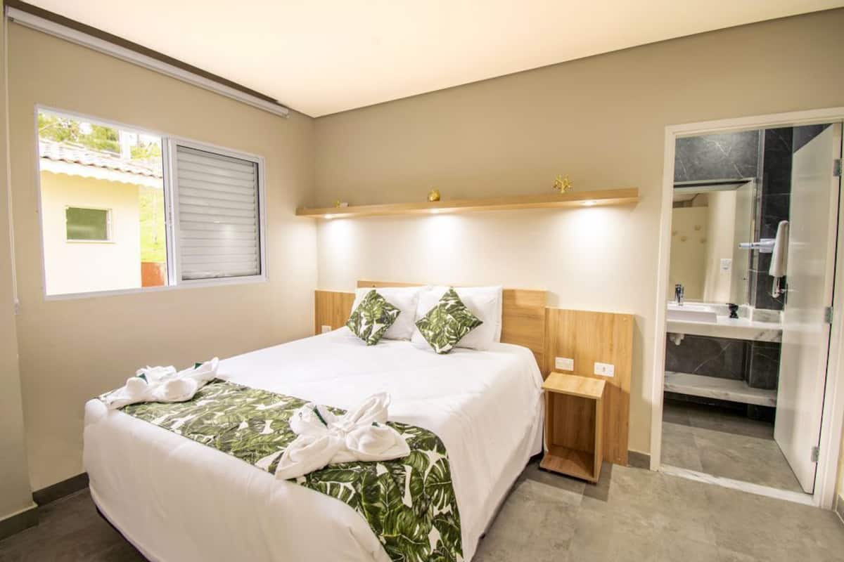 Une chambre d’hôtel comprenant un lit, une table de chevet, une commode, une salle de bain avec douche et une fenêtre avec stores.