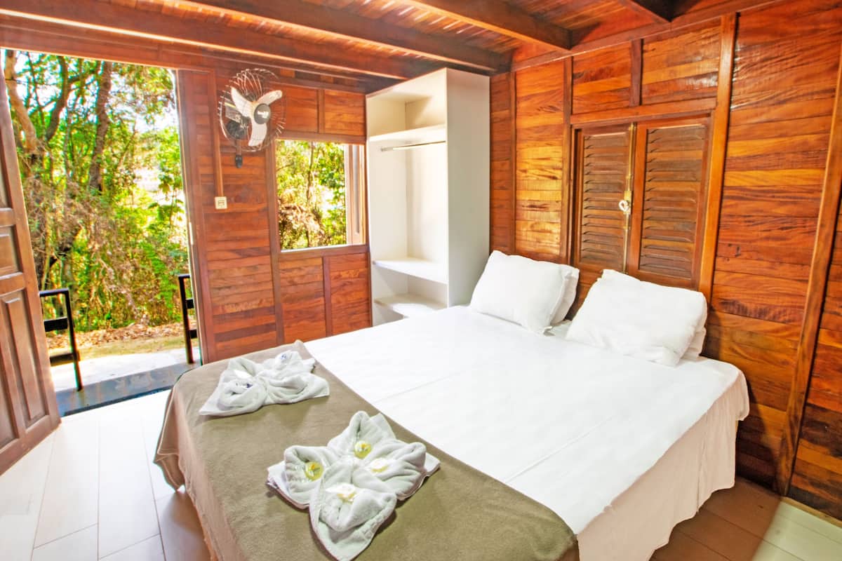 Une pièce en bois avec un lit, un ventilateur blanc fixé au mur et une fenêtre donnant sur un paysage verdoyant.