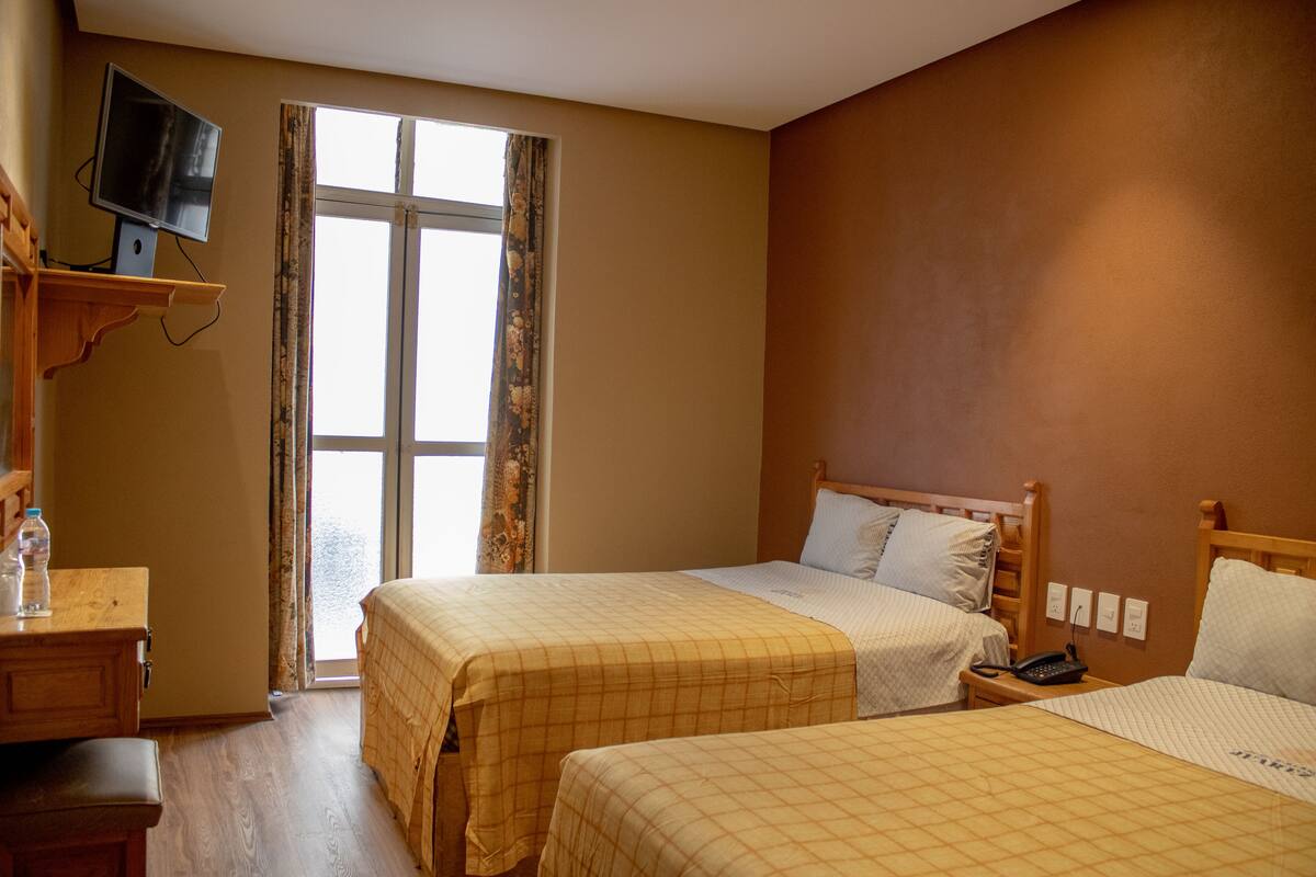 A hotel room with two beds, a TV on a shelf, a desk, and a window with curtains.