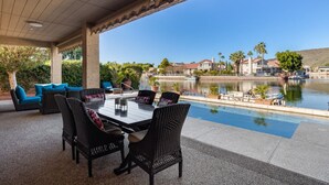 An outdoor patio with a dining table and chairs, overlooking a lake with houses and palm trees in the background.