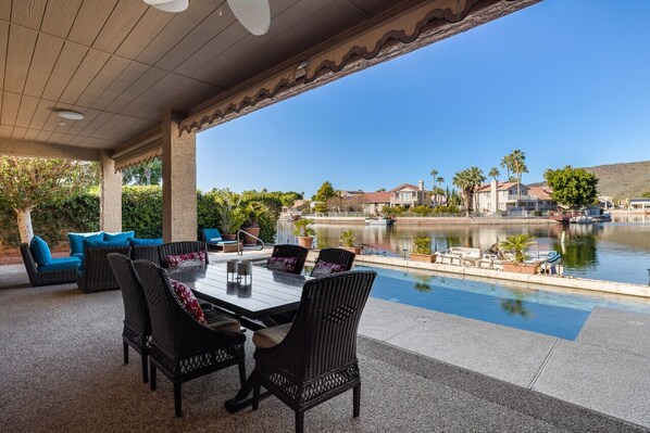 An outdoor patio with a dining table and chairs, overlooking a lake with houses and palm trees in the background.