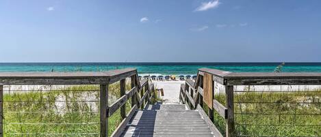 A wooden boardwalk leading to a beach with a clear view of the ocean and beach umbrellas in the distance.