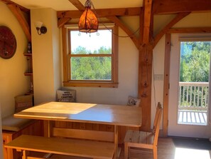 Cozy cabin-style room with a large window offering a view of the surrounding greenery, a wooden table, and a hanging light fixture.